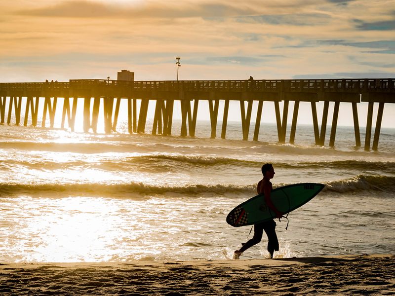 A surfer carrying a board along the shore at sunset, with a long pier behind