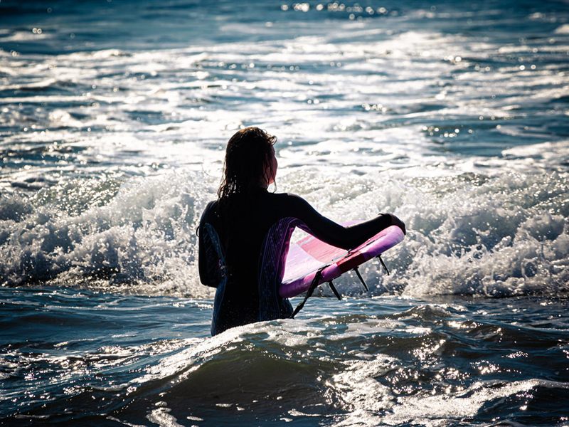 A surfer in a wetsuit wading into the whitewater with a pink board