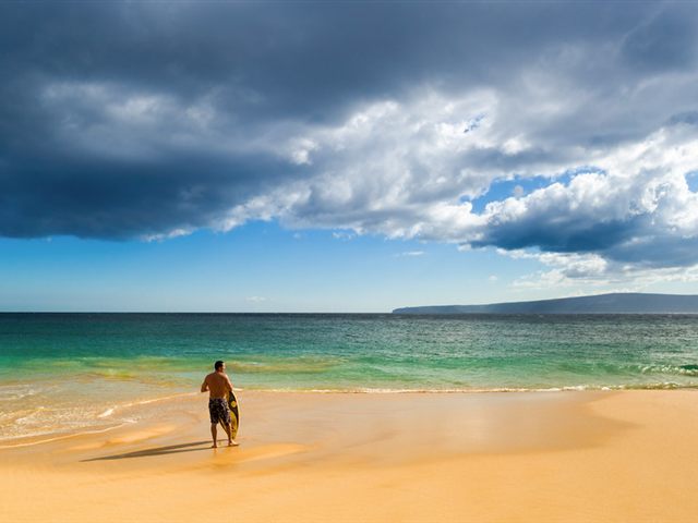 A man holding a small board at the water’s edge on a golden-sand beach, under dark clouds A man holding a small board at the water’s edge on a golden-sand beach, under dark clouds